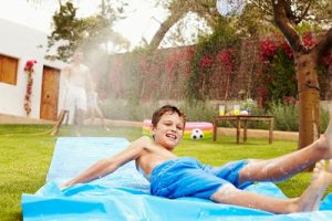 Father and kids having fun on a water slide in their garden using the hose